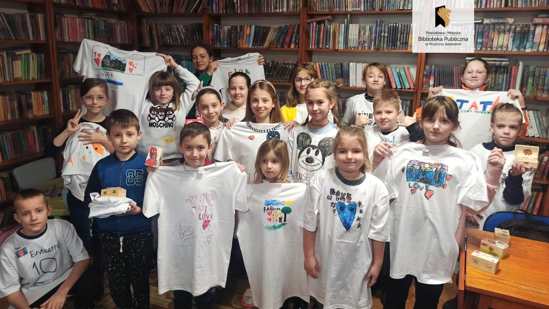 Osiemnaścioro dzieci stoi na tle regałów z książkami. Część z nich jest ubrana we własnoręcznie pomalowane koszulki, a część trzyma koszulki przed sobą. Kilkoro dzieci trzyma również ozdobione przez siebie szkatułki.