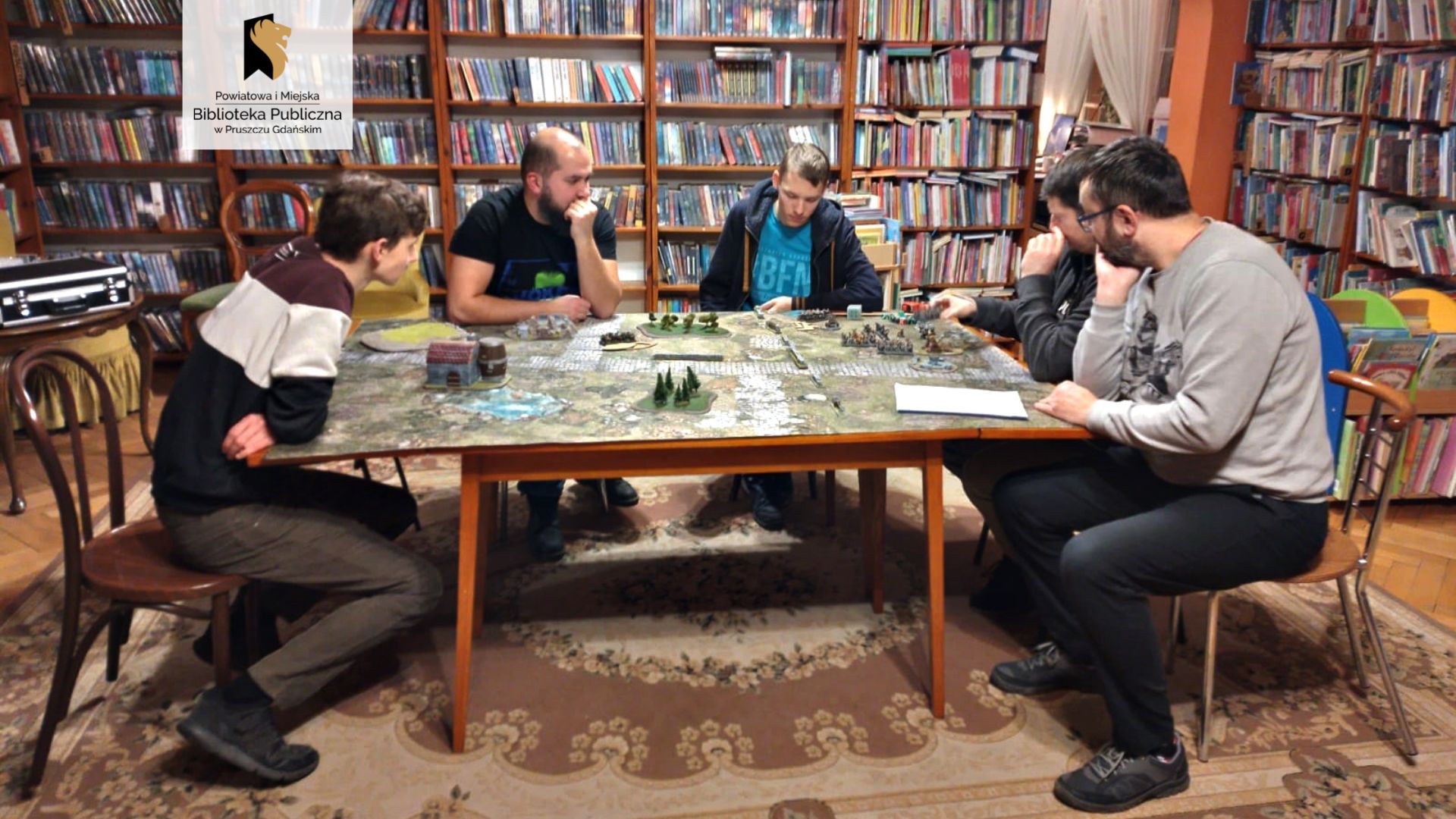 Sześć osób siedzi wokół dużego, prostokątnego stołu, na którym rozłożona jest wielka plansza do gry. Na planszy różne figurki postaci, drzew i budynków.