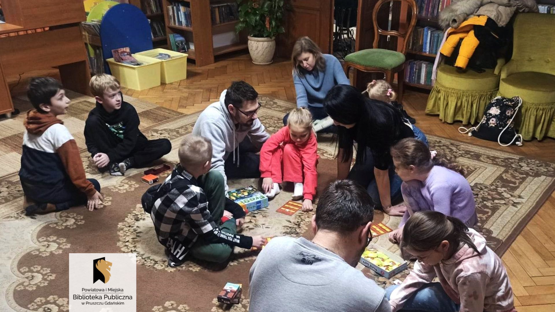 Siedmioro dzieci oraz czworo dorosłych siedzą na dywanie skupieni wokół kilku rozłożonych gier planszowych.