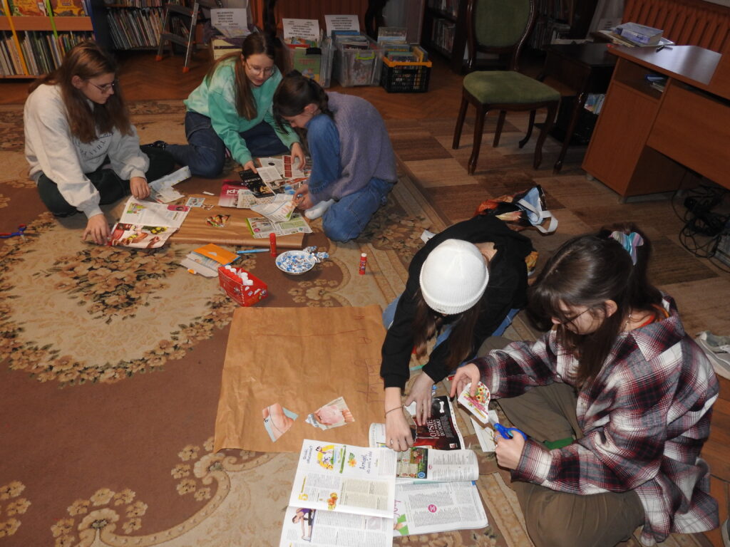 Pięcioro dzieci siedzi lub klęczy na dywanie. Są podzieleni na dwie grupy. Przed każdą grupą arkusz szarego papieru oraz liczne czasopisma. Dzieci wycinają wybrane ilustracje z gazet.