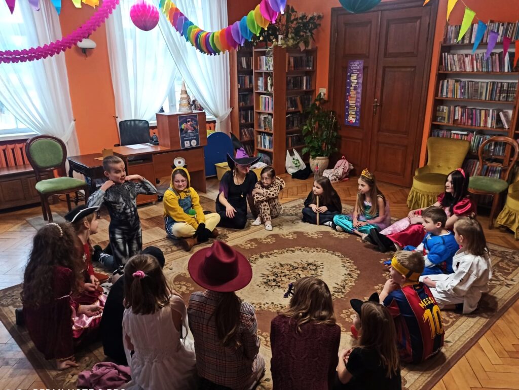 Duża grupa dzieci, bibliotekarka oraz wolontariuszka siedzą na dywanie w kole. Wszyscy ubrani są w stroje karnawałowe np. czarownicy, wilkołaka, kota, detektywa, księżniczki, superbohaterów… Pod sufitem zawieszone kolorowe girlandy.