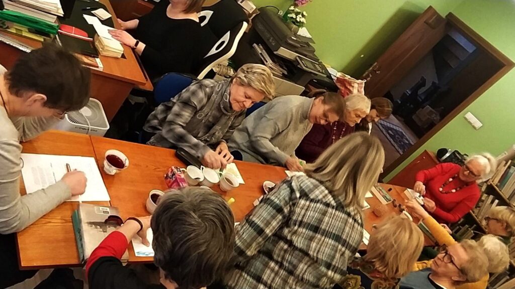Kilka kobiet siedzi wokół długiego, prostokątnego stołu. Na stole filiżanki z herbatą, kartki, mazaki, książki. Panie rysują coś na kartkach.