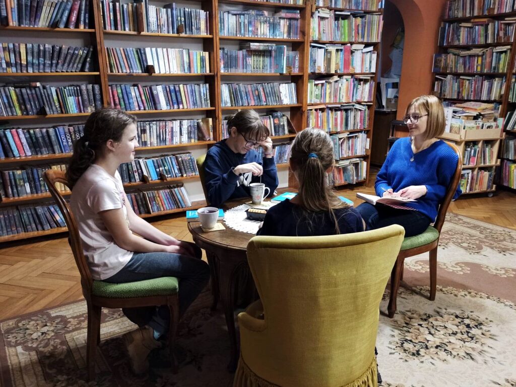 Trzy nastolatki oraz bibliotekarka siedzą na krzesłach i fotelach wokół stołu. Na stole książki, kubki z herbatą, pudełko ciastek.