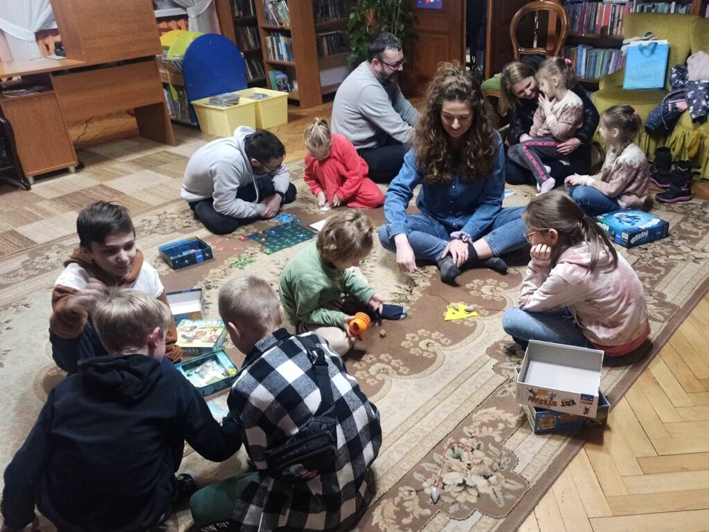 Ośmioro dzieci oraz czworo dorosłych siedzą na dywanie skupieni wokół kilku rozłożonych gier planszowych.