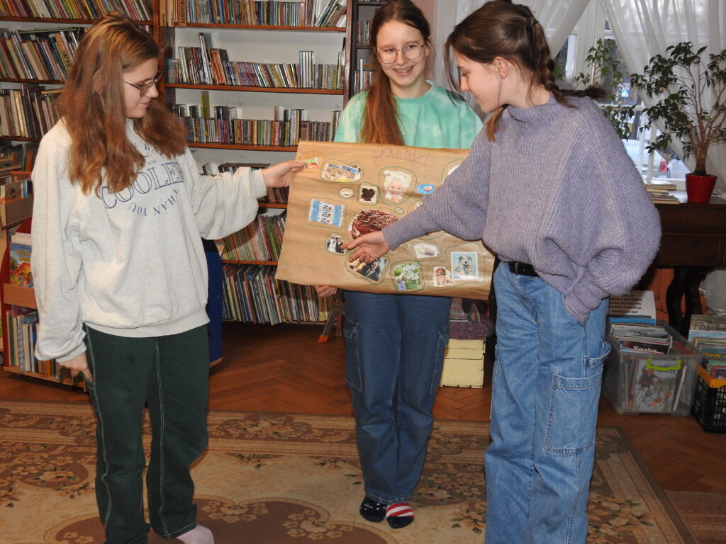 Trzy dziewczyny stoją. Dwie z nich trzymają arkusz szarego papieru z ponaklejanymi fragmentami czasopism. Jedna wskazuje palcem na poszczególne zdjęcia.
