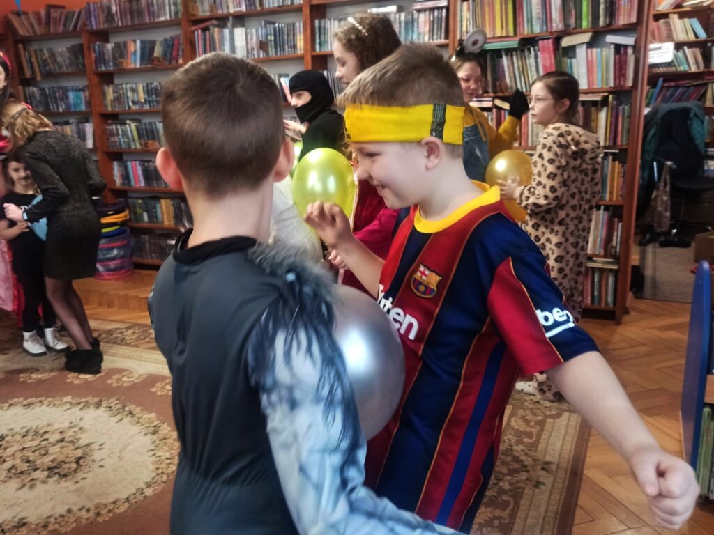 Dwoje chłopców ubranych w stroje wilkołaka i piłkarza tańczy z balonem pomiędzy brzuchami. W tle inne pary tańczące w ten sam sposób.