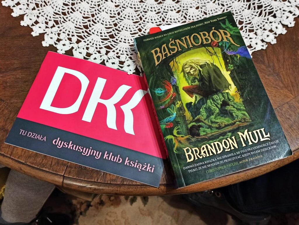 Na stole leży logo Dyskusyjnego Klubu Książki oraz książka pt. Baśniobór.