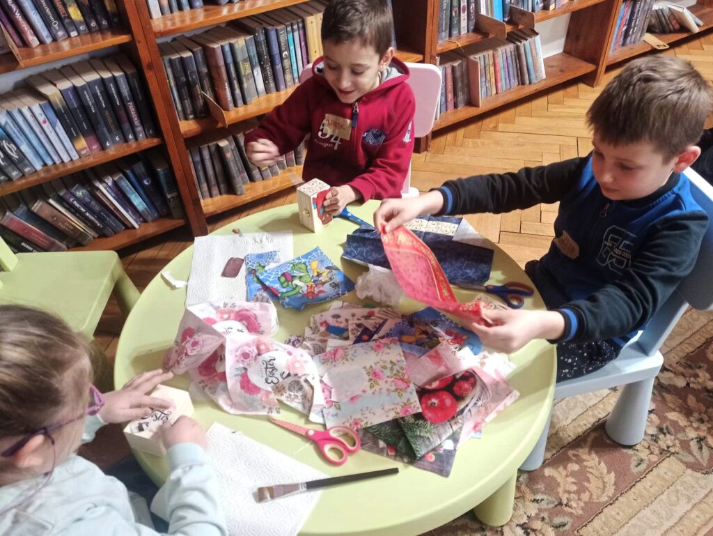 Troje dzieci siedzi przy małym, okrągłym stoliku, na którym znajdują się serwetki, nożyczki, pędzle, pudełko z klejem. Jedno dziecko wybiera serwetki, a dwoje nakleja fragmenty serwetek na szkatułki.