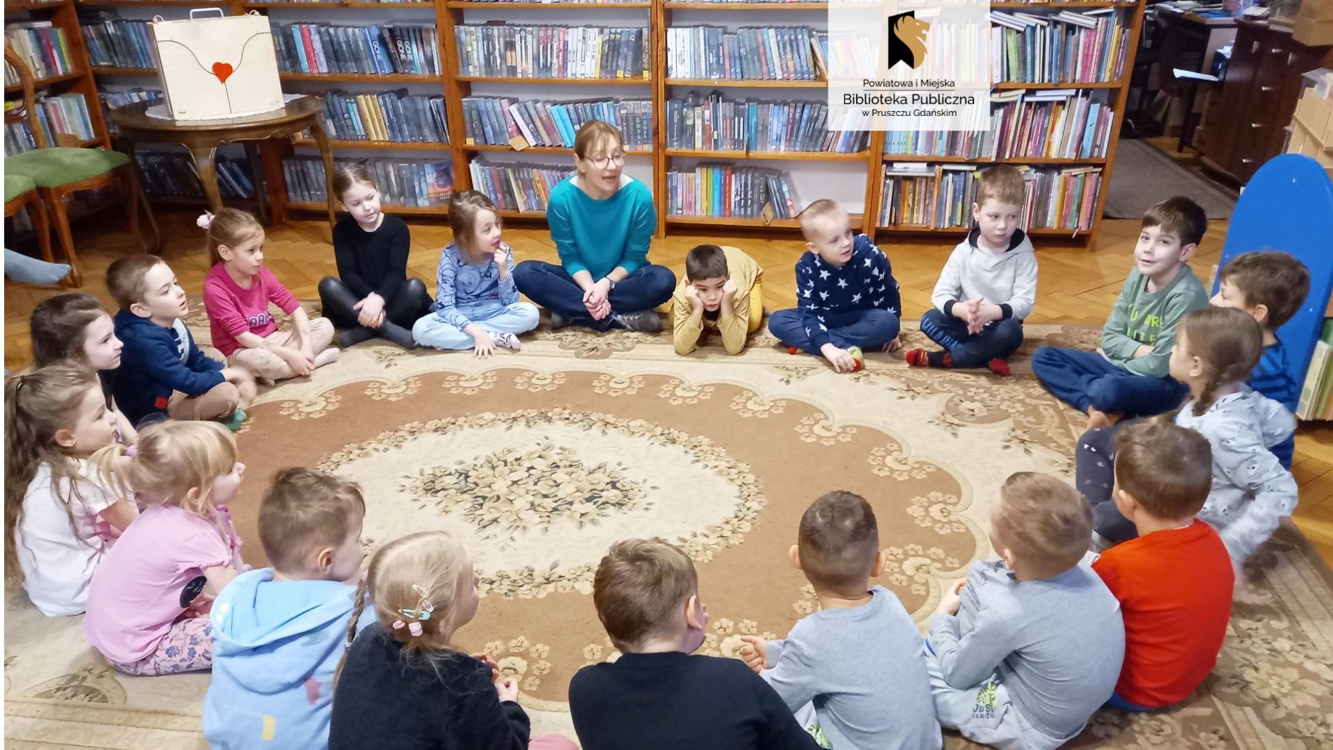 Na dywanie w kole siedzą po turecku dzieci. Pomiędzy nimi bibliotekarka. W rogu sali stoi okrągły brązowy stół a na nim drewniany teatrzyk.
