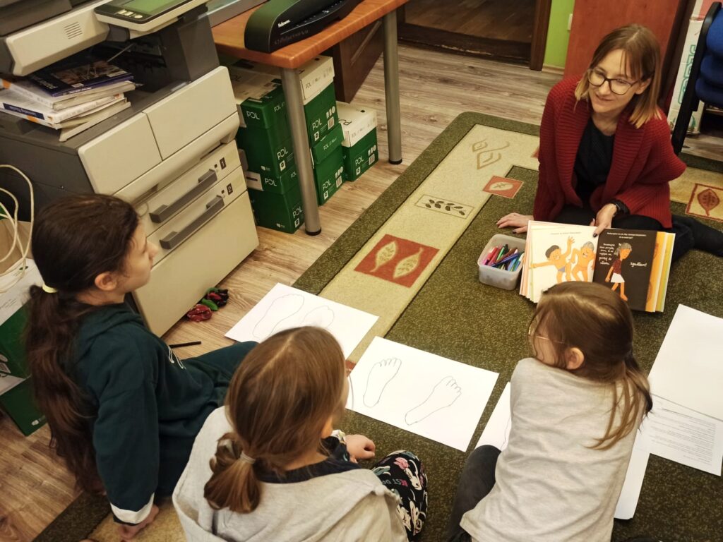Trzy dziewczynki siedzą naprzeciw bibliotekarki, która trzyma książkę otwartą na ilustracji przedstawiającej troje dzieci. Pomiędzy nimi, na dywanie leżą kartki z odrysowanymi stopami.