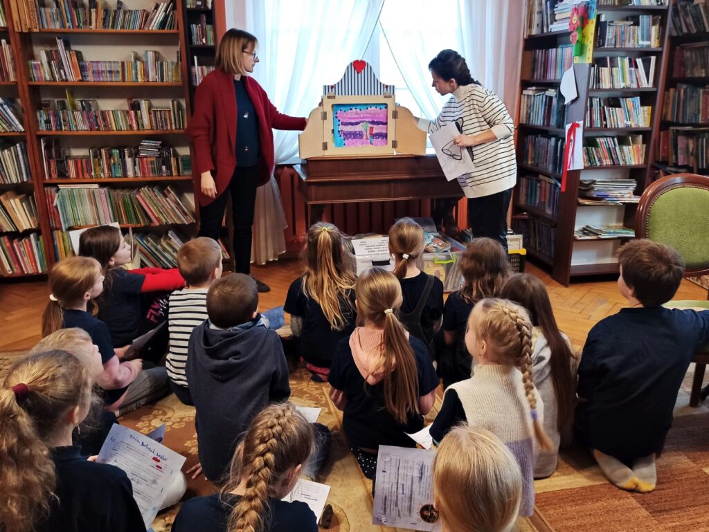 Bibliotekarka, Joanna Łabasiewicz, oraz nauczycielka, Katarzyna Ciepielewska, stoją przy sekretarzyku, na którym stoi otwarty teatrzyk kamishibai. Dotykają karty w teatrzyku. Naprzeciw siedzi grupa kilkunastu dzieci. 