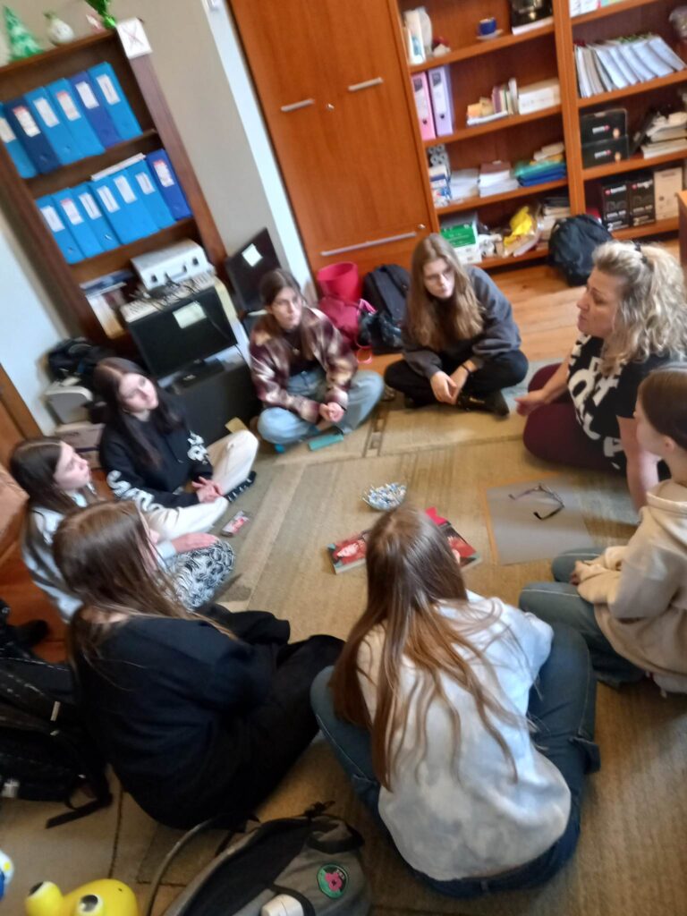 7 dziewczyn z młodzieżowego Dyskusyjnego Klubu Książki oraz moderatorka klubu siedzą w okręgu na dywanie i rozmawiają o książce. Po środku stoi miska z cukierkami oraz leżą 3 książki i logotyp klubu.