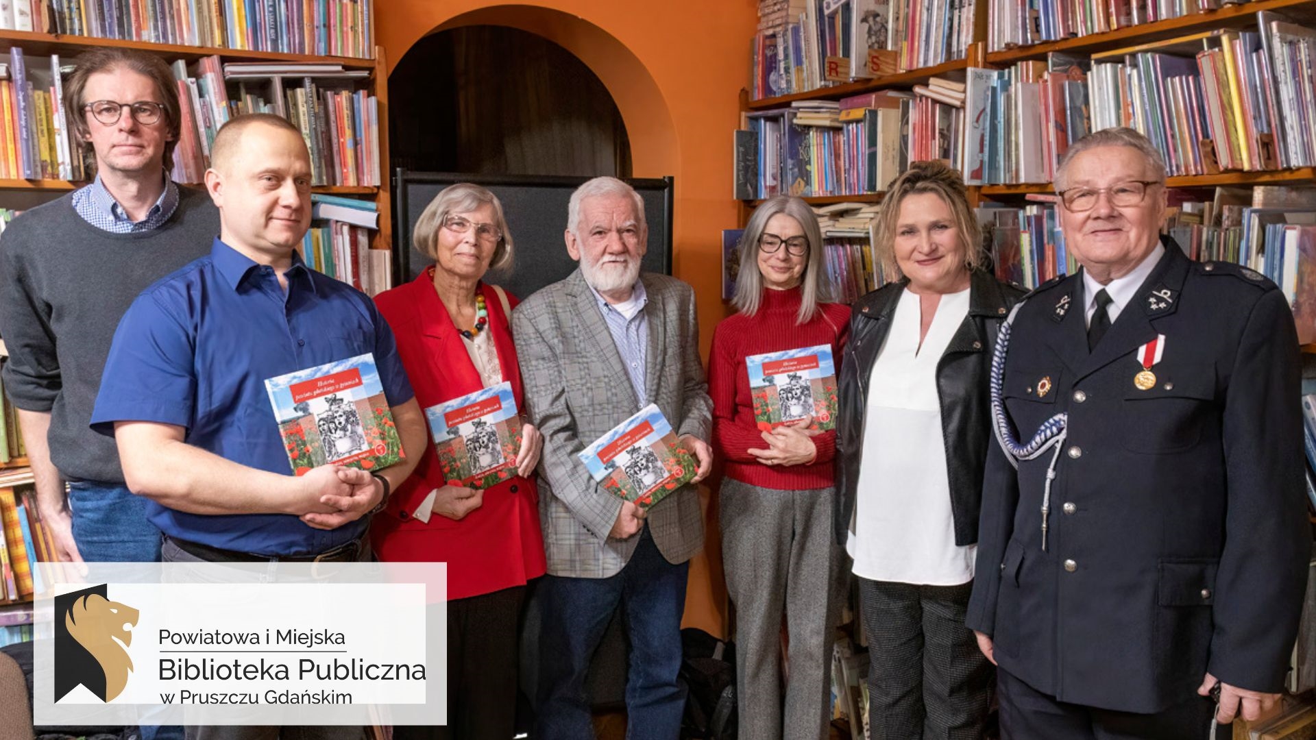 7 osób pozuje do zdjęcia w bibliotece. 4 osoby trzymaja w rękach promowaną podczas spotkania książkę pt.Historia powiatu gdańskiego w pytaniach.