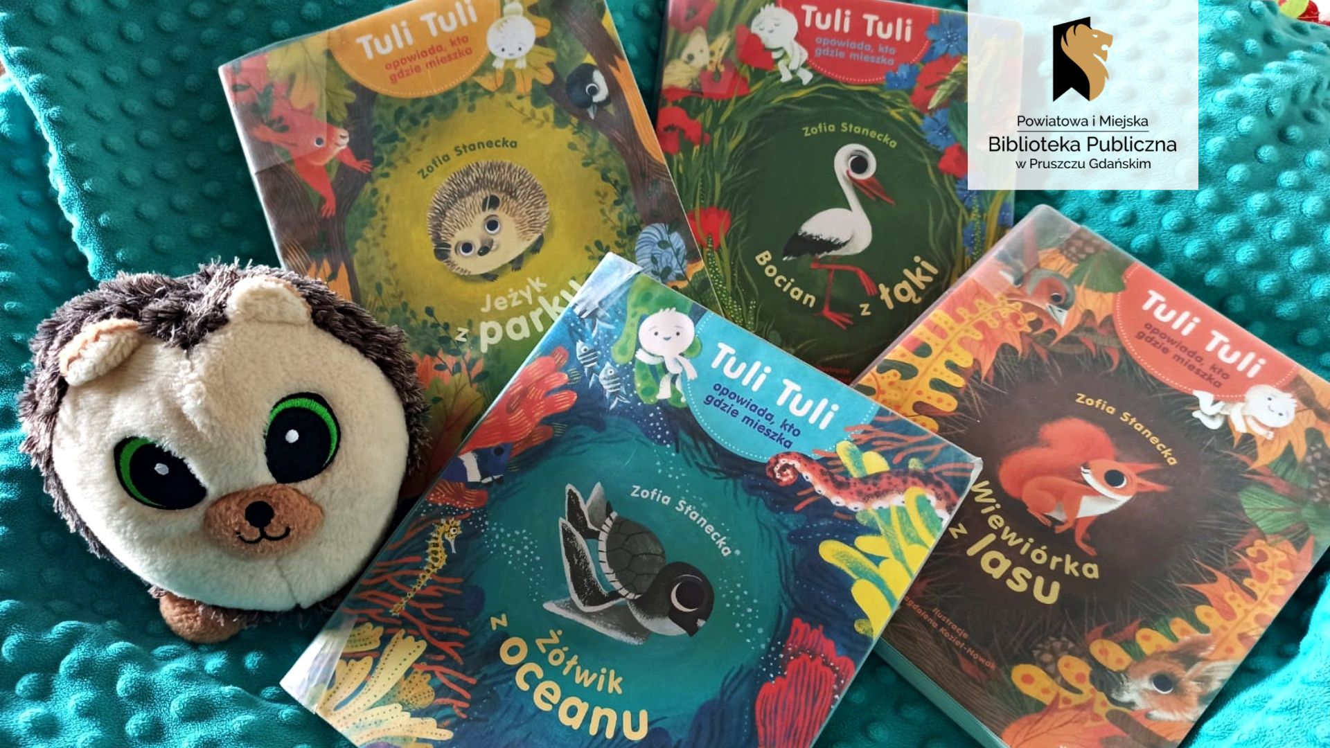 Cztery książki z serii Tuli Tuli opowiada, kto gdzie mieszka leżą na niebieskim kocu. Na okładkach ilustracje zwierząt: jeża, bociana, wiewiórki, żółwia. Obok książek - pluszowy jeż.