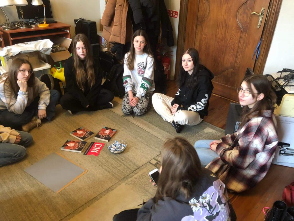 6 dziewczyn z młodzieżowego Dyskusyjnego Klubu Książki siedzi na dywanie w okręgu. Po środku stoi miska z cukierkami oraz leżą 3 książki i logotyp klubu.