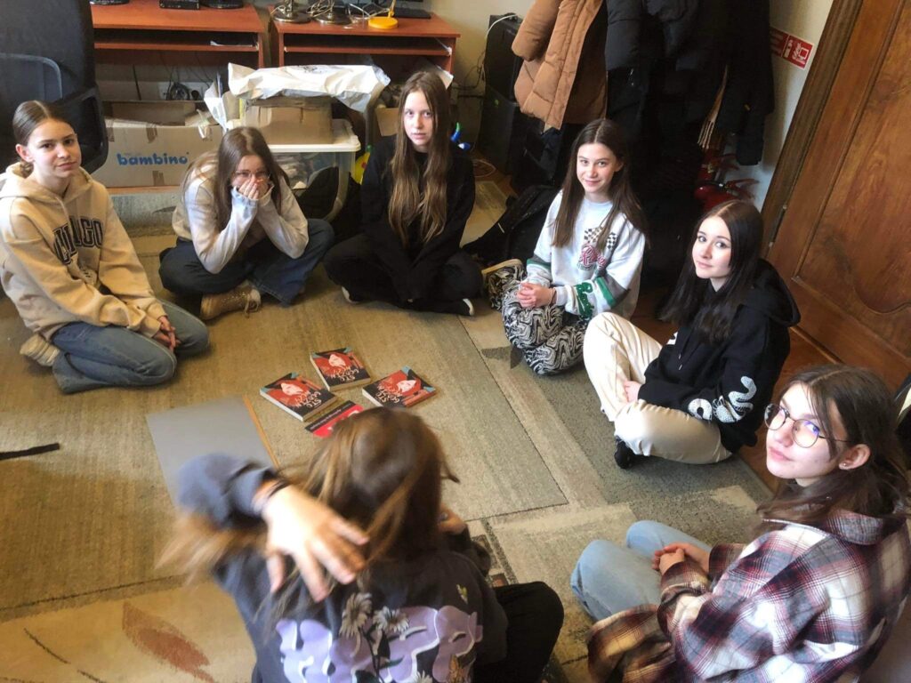 6 dziewczyn z młodzieżowego Dyskusyjnego Klubu Książki siedzi na dywanie w okręgu. Po środku stoi miska z cukierkami oraz leżą 3 książki i logotyp klubu.