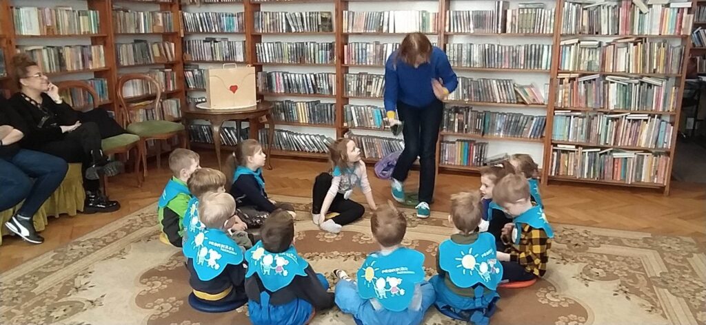 Grupa dzieci przedszkolnych siedzi na dywanie w pomieszczeniu bibliotecznym. Dzieci siedza w okręgu i patrzą na bibliotekarkę, która się nad nimi pochyla.