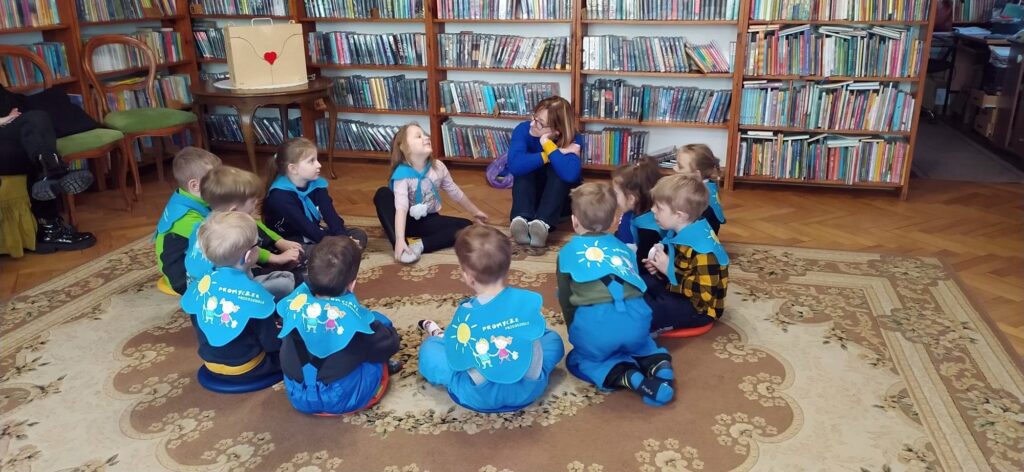 Grupa dzieci przedszkolnych siedzi na dywanie w pomieszczeniu bibliotecznym. Dzieci siedza w okręgu i patrzą na bibliotekarkę, która do nich mówi.
