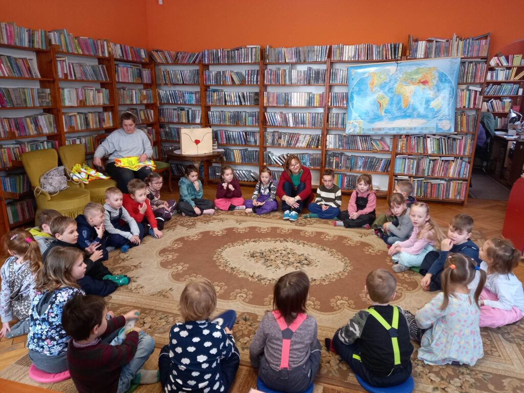 Grupa dzieci i bibliotekarka siedzą w bibliotece na dywanie w okręgu. Z tyłu, na regale z książkami zawieszona jest mapa.