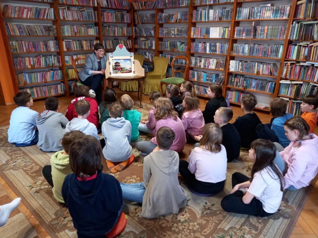 Duża grupa dzieci siedzi na dywanie w bibliotece. Dzieci zwrócone są w stronę bibliotekarki, która czyta im bajkę o Słowiku H.CH. Andersena. Bajka zaprezentowana jest w postaci teatrzyku kamishibai.