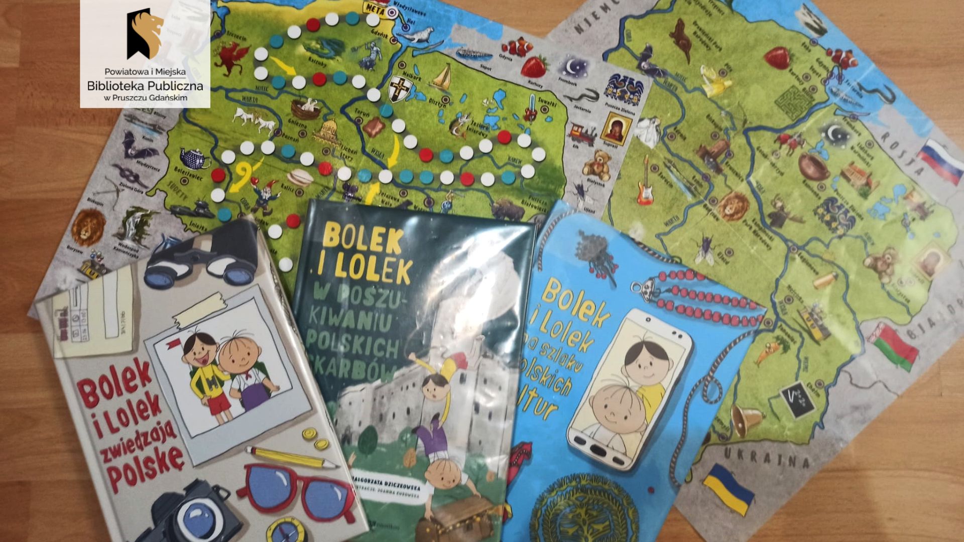 Trzy książki o Bolku i Lolku oraz dwie mapy Polski leżą na podłodze. Mapy są przeznaczone dla dzieci, nadrukowane są nich liczne ilustracje przedstawiające najważniejsze atrakcje określonych miejsc.