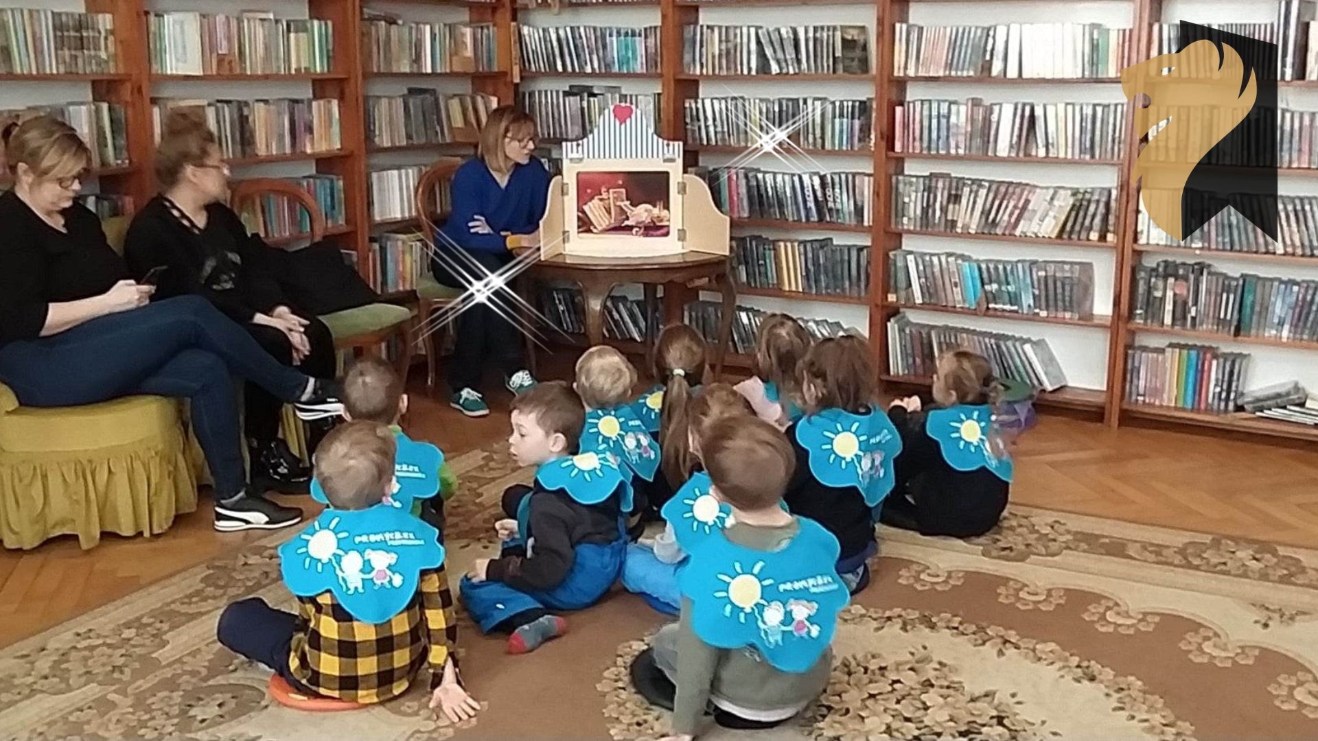 Grupa dzieci przedszkolnych siedzi na dywanie zwrócona w stronę bibliotekarki, która czyta historię z teatrzyku kamishibai.