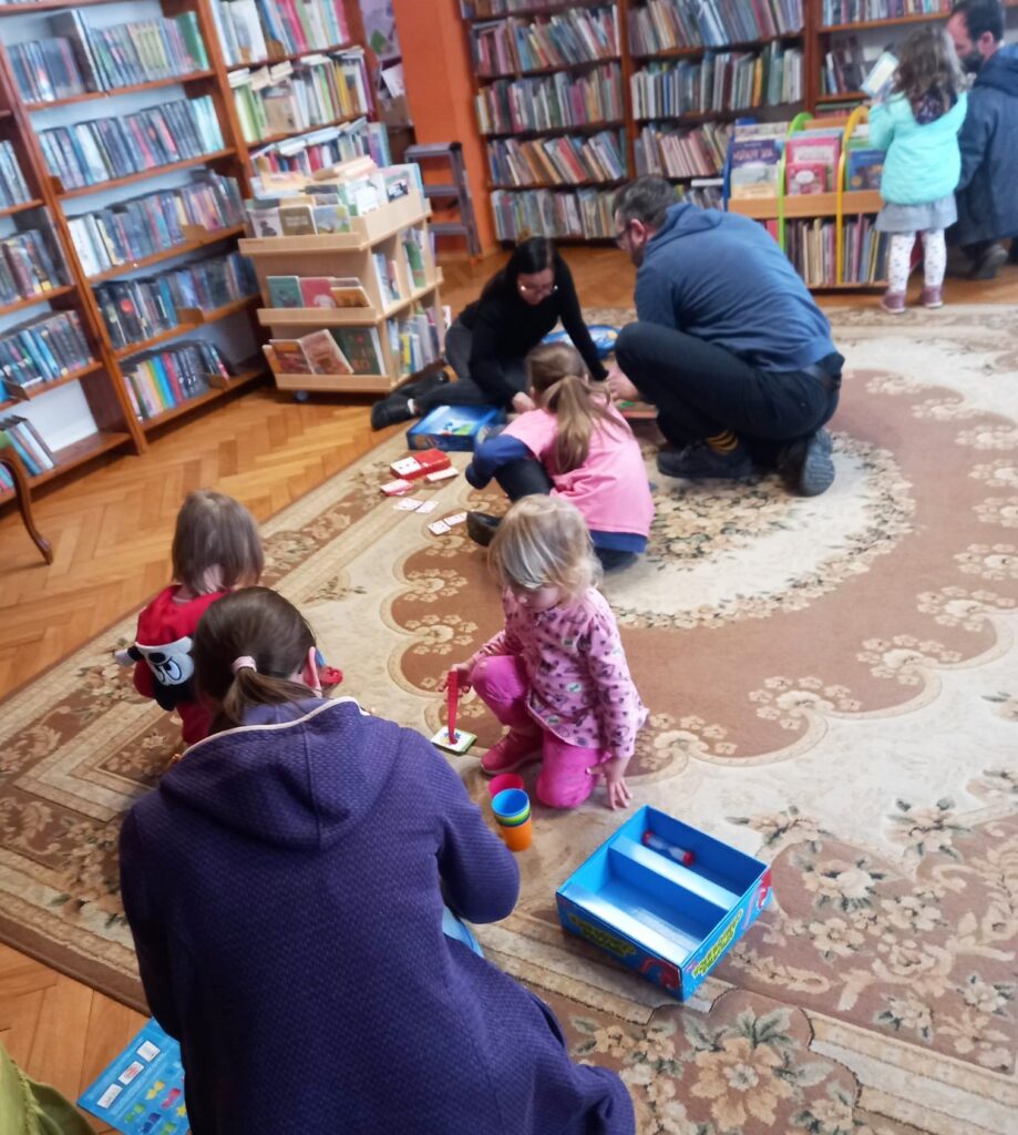 Na dywanie siedzą dorośli i  dzieci. Przed nimi są rozłożone różne gry planszowe.