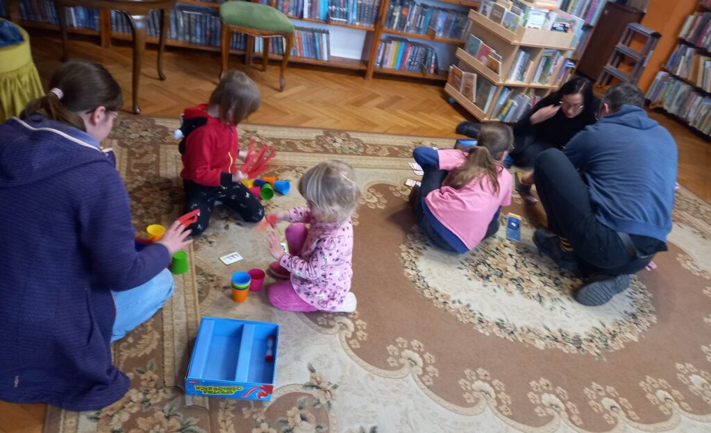 Na dywanie siedzą dorośli i  dzieci. Przed nimi są rozłożone są różne gry planszowe.