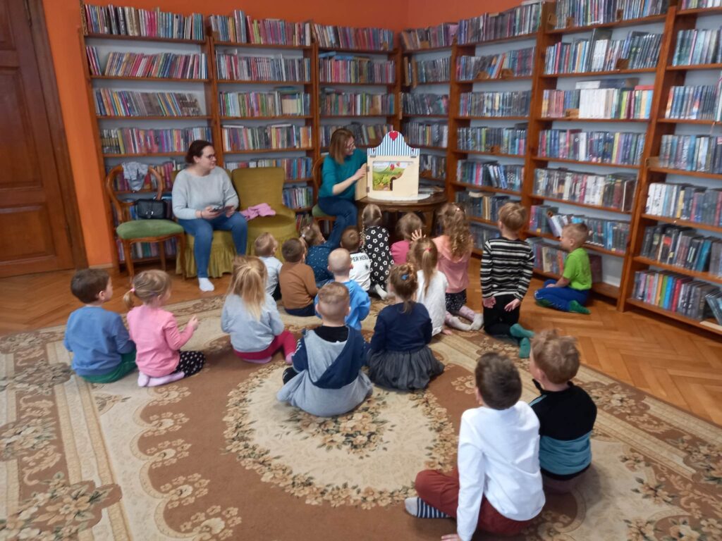 Dzieci siedzą na dywanie zwrócone w stronę bibliotekarki, która czyta Magiczną gąsienice. Książeczka przedstawiona jest za pomocą teatrzyku kamishibai.