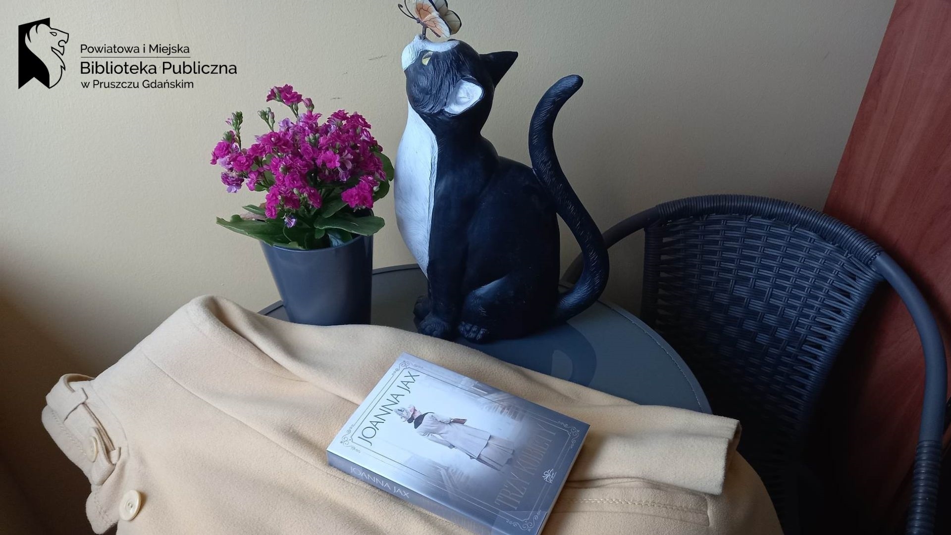 Na stoliku na jasnym płaszczu leży książka Joanny Jax pt.Trzy kobiety. Obok, w doniczce stoi kwiat oraz figurka kota z motylem na nosie.