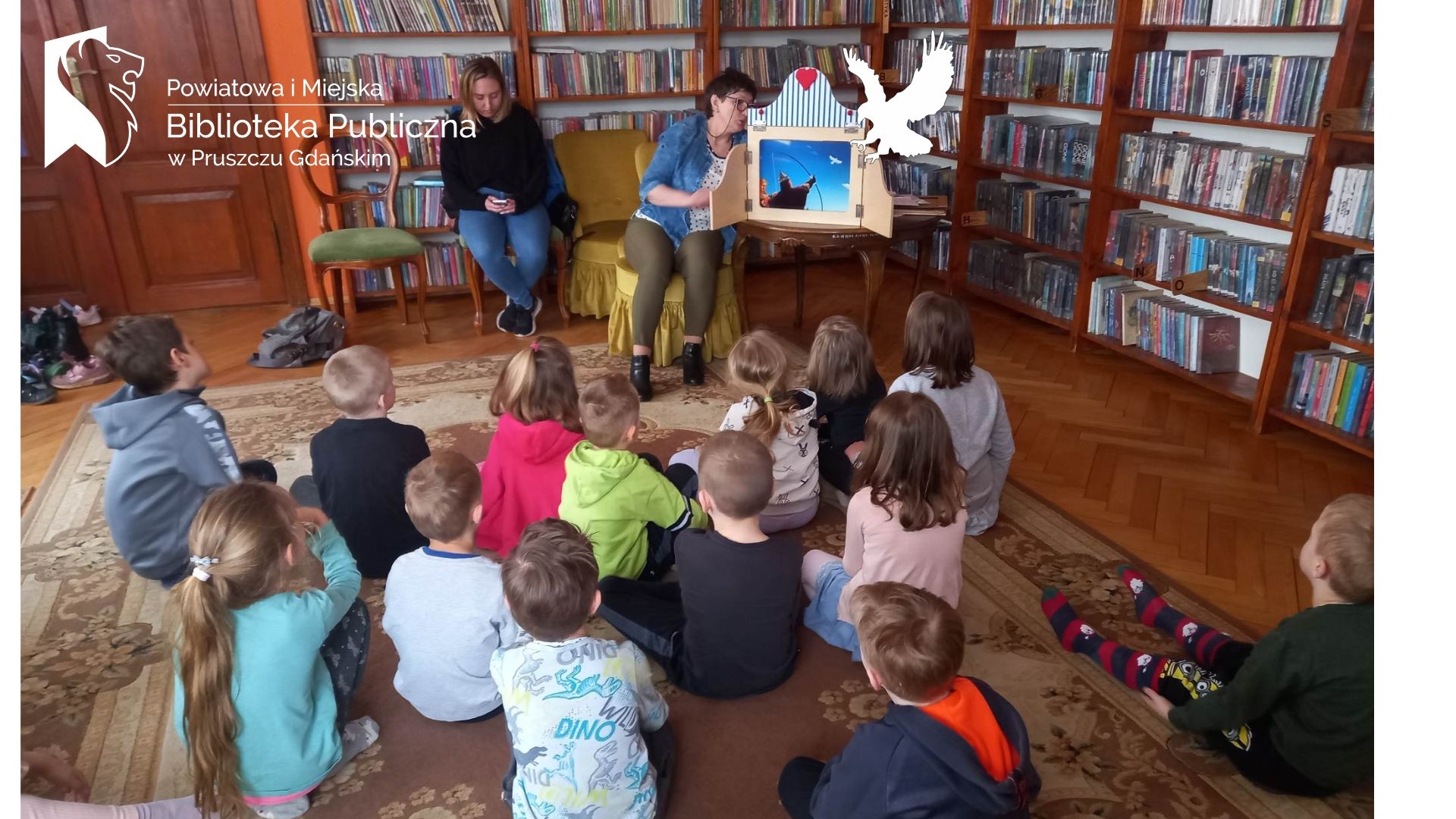 Grupa dzieci siedzi na dywanie i słucha Legendy o Lechu, Czechu i Rusie, która prezentuje im bibliotekarka przy pomocy teatrzyku kamishibai.
