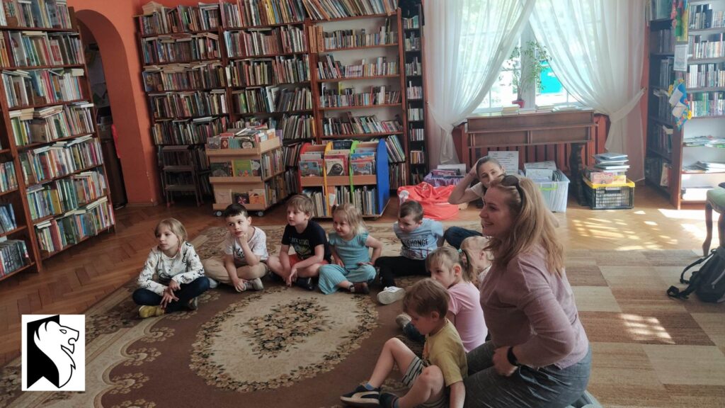 1.	Dzieci wraz z opiekunkami siedzą w półkole na podłodze biblioteki. Za nimi regały z książkami. 