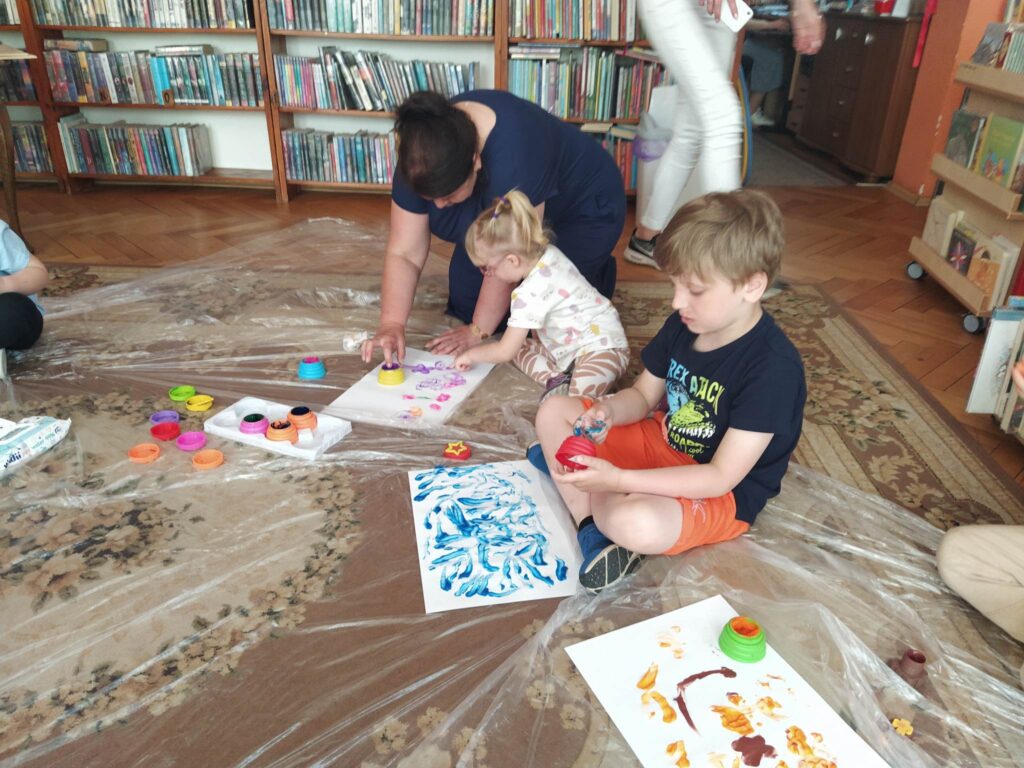 2.	Chłopiec i dziewczynka wraz z Panią Opiekunką siedzą na podłodze i malują farbami palcami.