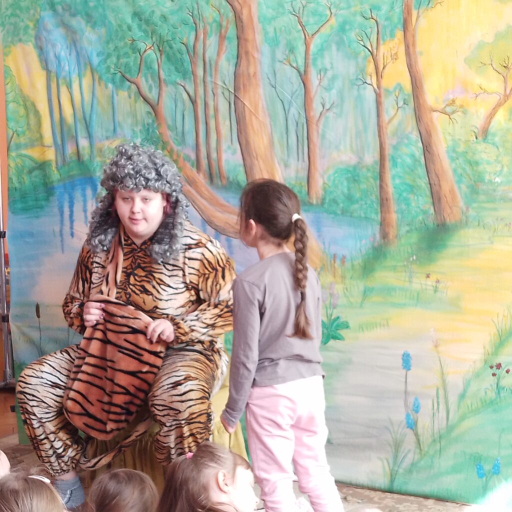 Scena przedstawiająca krajobraz: jezioro, zielone drzewa, kwiaty oraz dróżkę. Na scenie 2 aktorów.  Na scenie siedzi aktor ubrany w strój tygrysa, obok niego stoi dziewczynka z widowni.