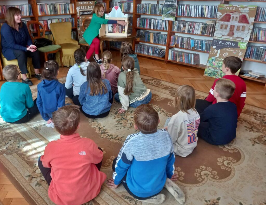 Na dywanie siedzą dzieci. Przed nimi siedzi bibliotekarka, która prezentuje baśń Andersena pt. Słowik za pomocą teatrzyku kamishibai.