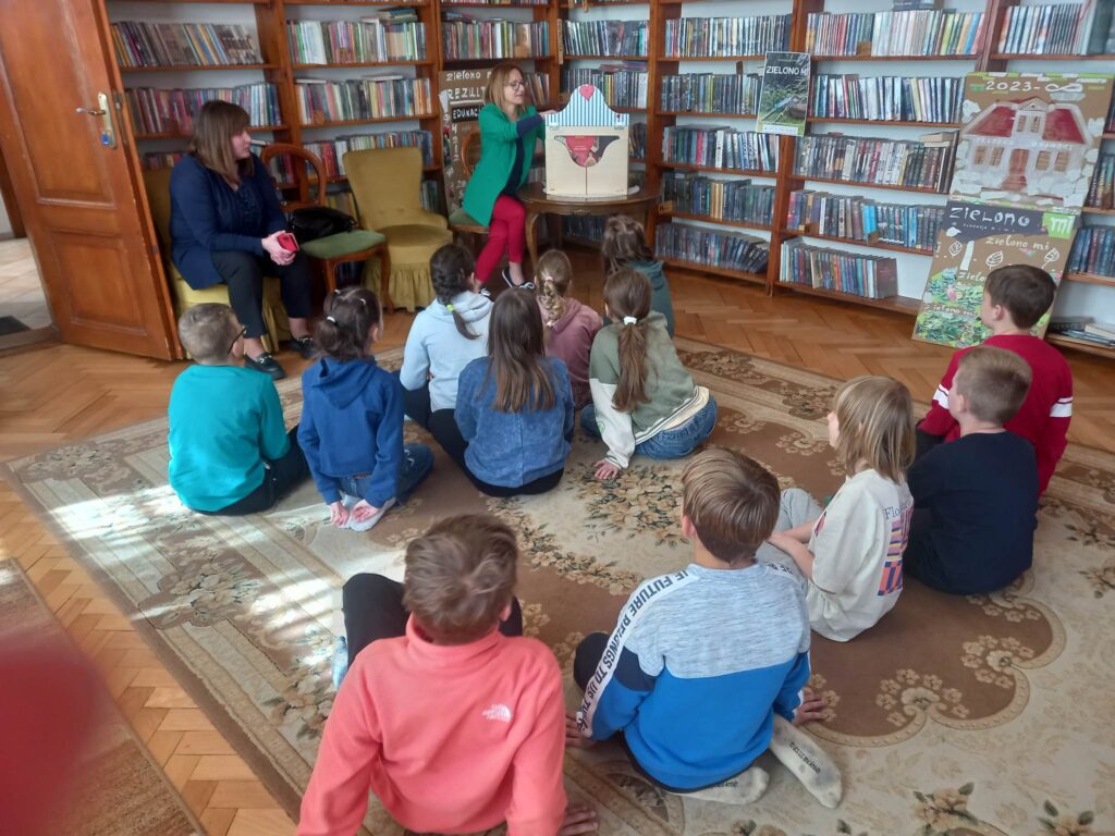 Na dywanie siedzą dzieci. Przed nimi siedzi bibliotekarka, która otwiera teatrzyk kamishibai.
