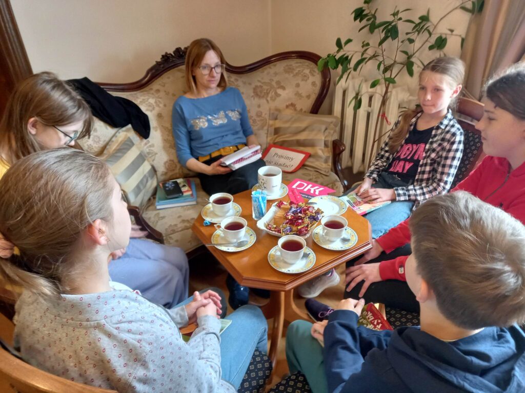 Moderatorka Dyskusyjnego Klubu Książki Joanna Łabasiewicz oraz 5 dzieci siedzą przy stoliku i rozmawiają o książce.