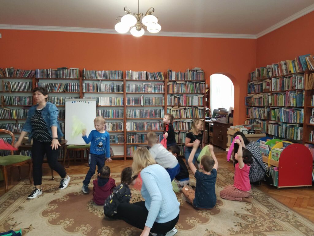 Bibliotekarka oraz część dzieci bawią się w motyle i biegają po sali. Część dzieci siedzi na dywanie.