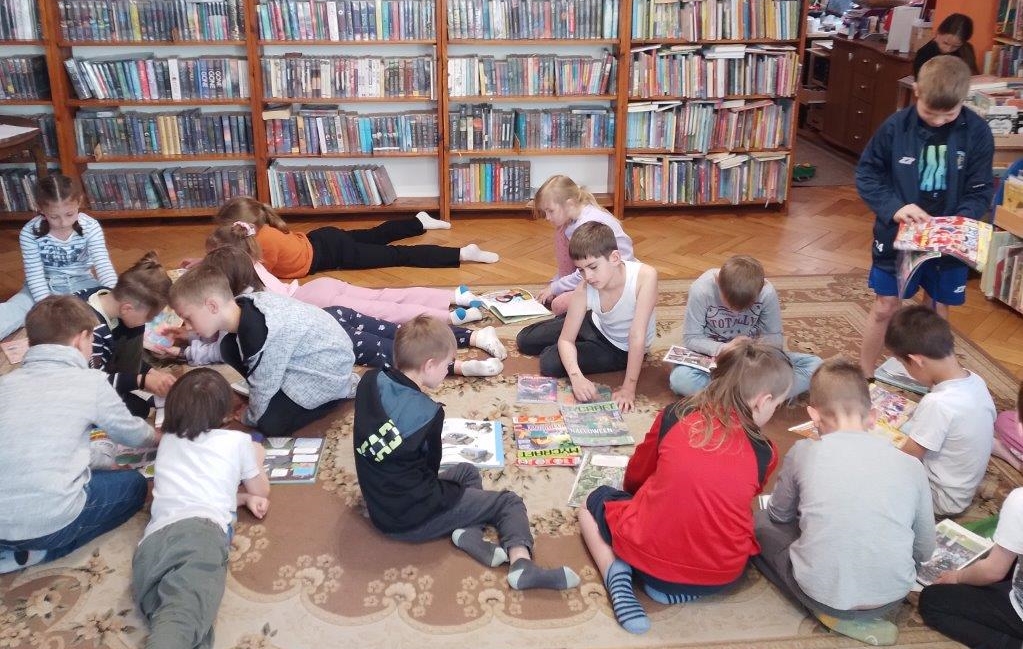 Na dywanie w bibliotece siedzą na dywanie dzieci i oglądają książki.
