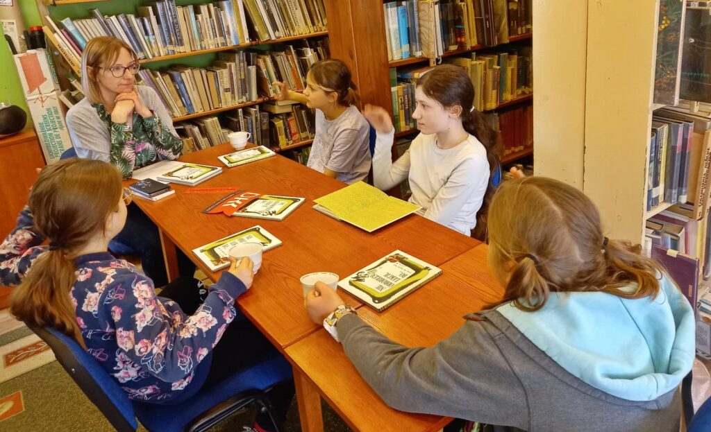 Czworo dzieci z Dyskusyjnego Klubu Ksiąski oraz moderatorka siedzą przy stole. Na stole leżą książki oraz logotyp klubu. Jedno z dzieci zgłasza się.
