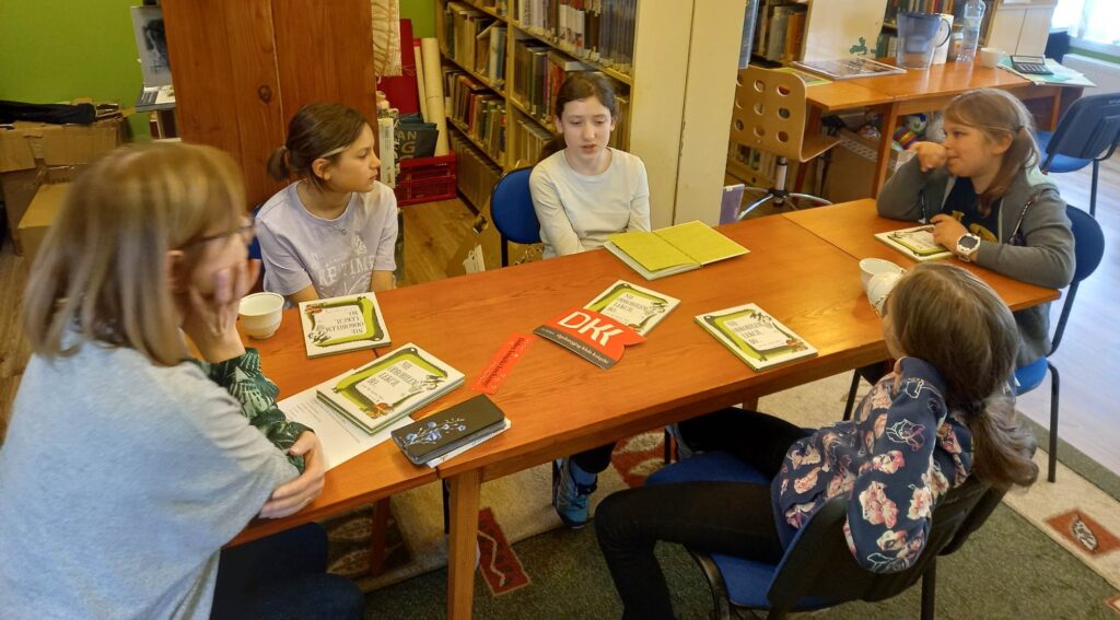 Czworo dzieci z Dyskusyjnego Klubu Ksiąski oraz moderatorka siedzą przy stole. Na stole leżą książki oraz logotyp klubu.