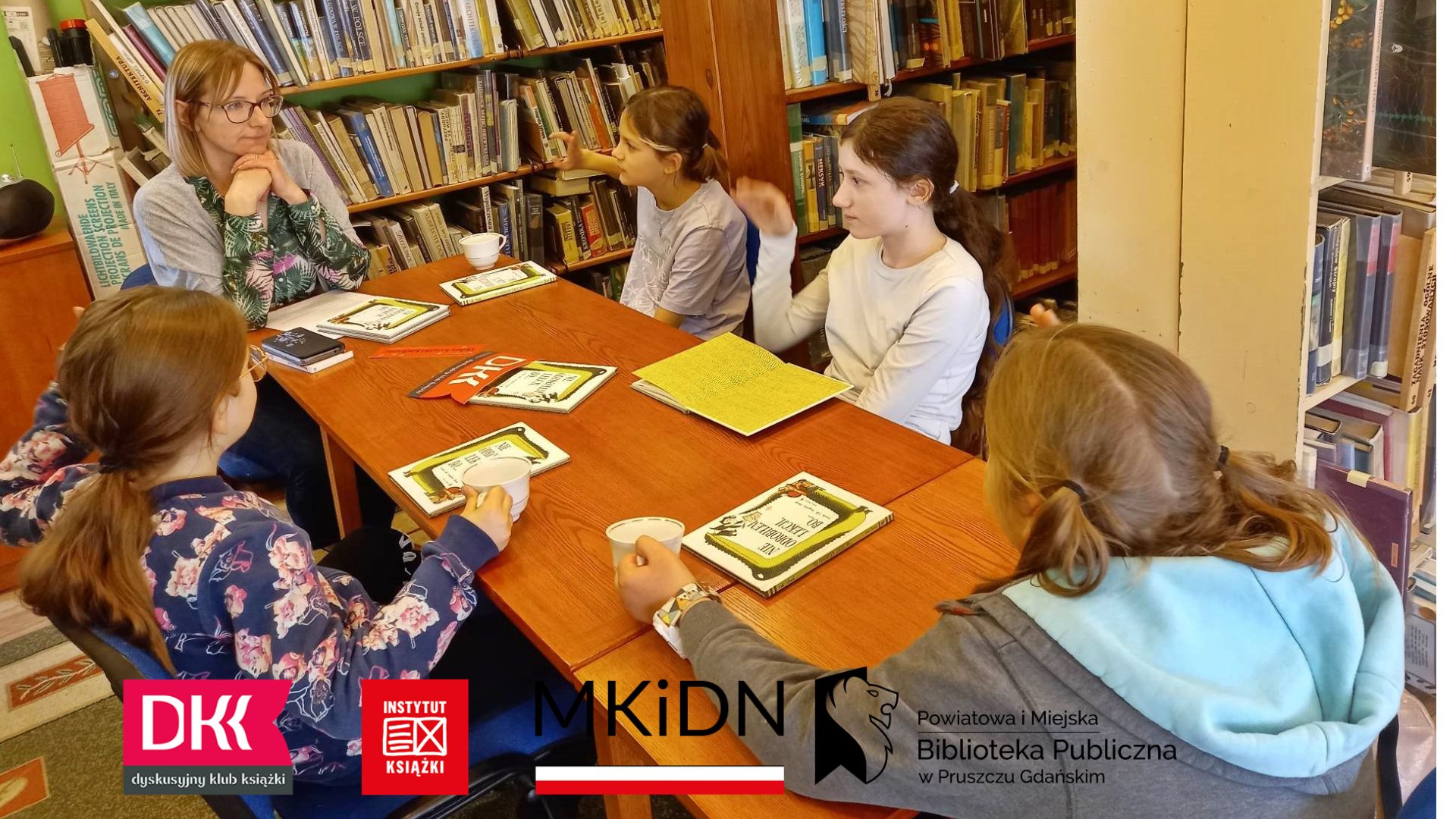 Czworo dzieci z Dyskusyjnego Klubu Ksiąski oraz moderatorka siedzą przy stole. Na stole leżą książki oraz logotyp klubu.
