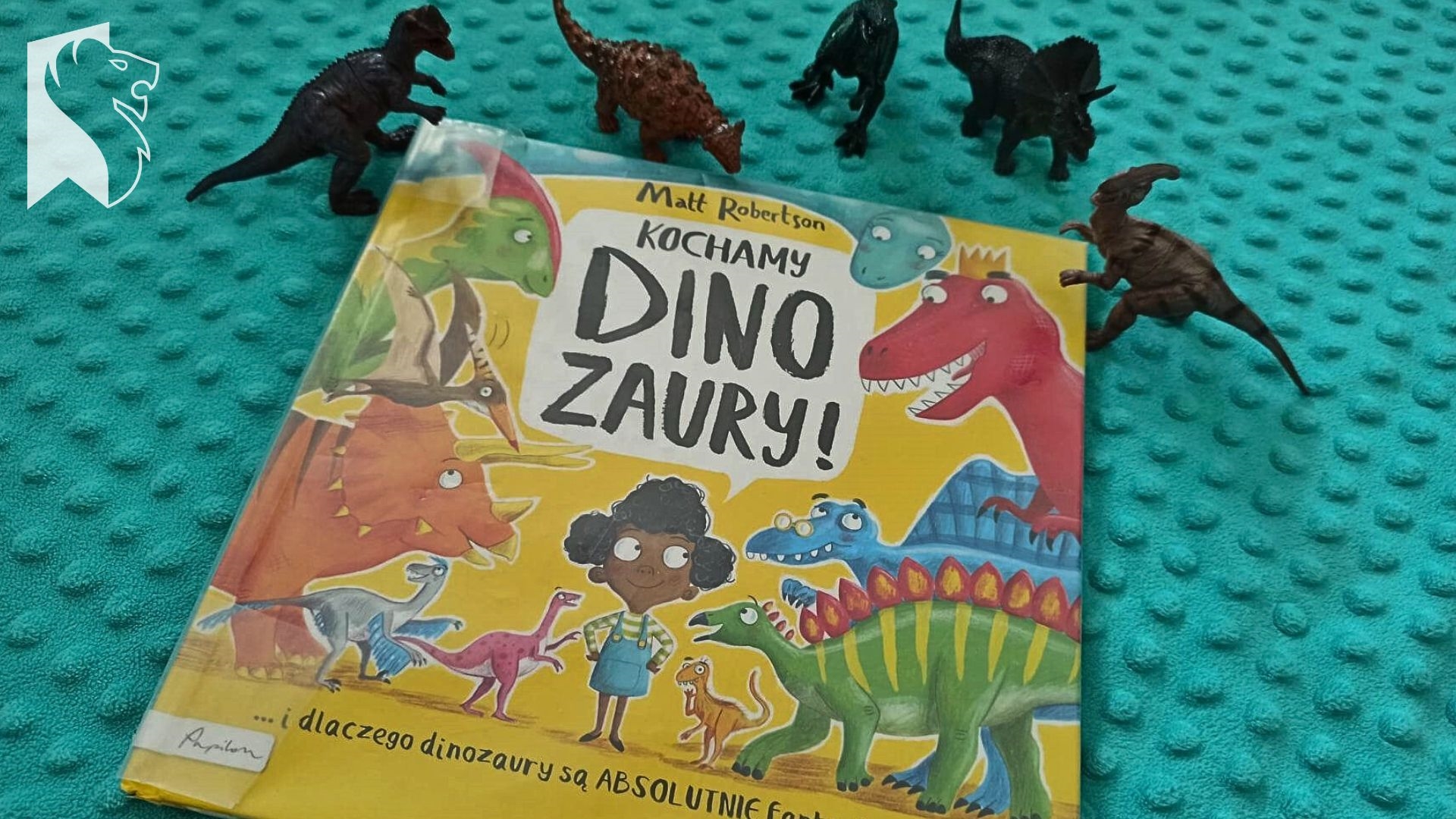 Na niebieskim kocyku książka Matta Robertsona pt. Kochamy dinozaury. Nad książką 5 małych figurek dinozaura.