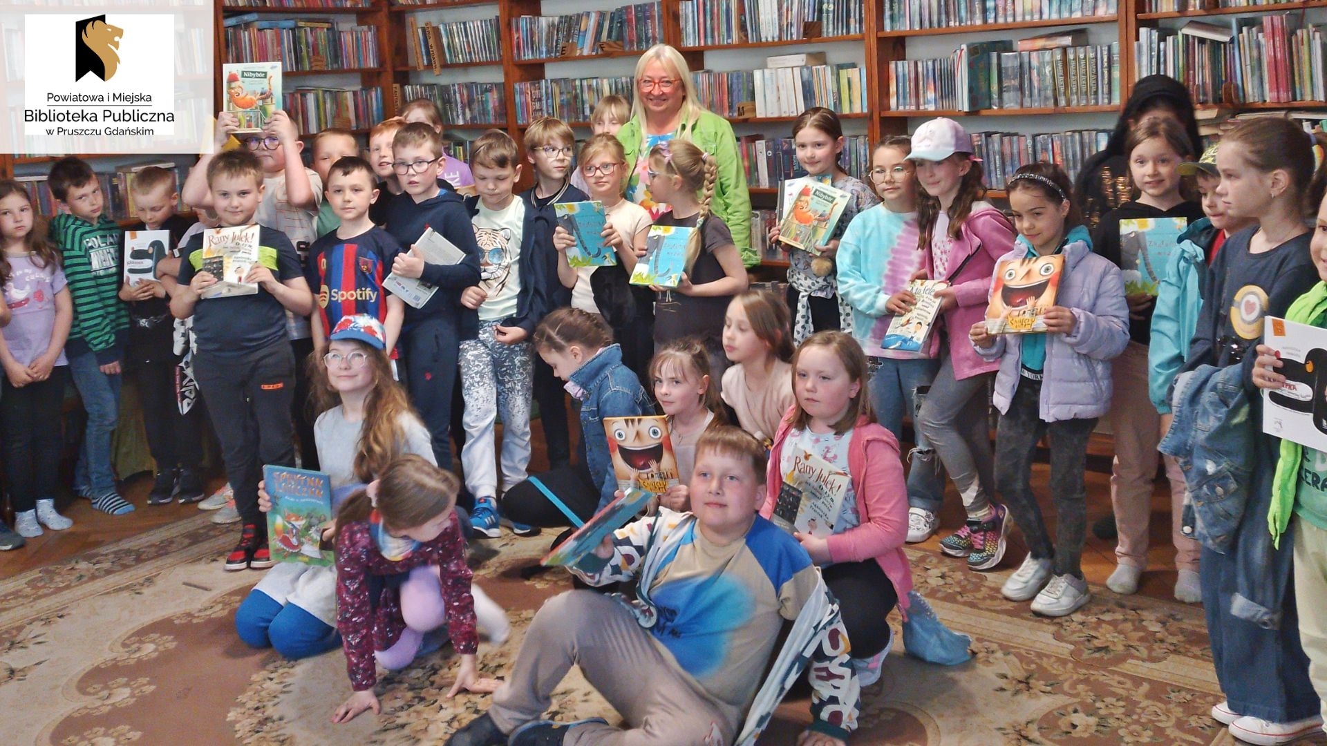 Pisarka Agnieszka Frączek pozuje do zdjęcia z dziećmi obecnymi podczas spotkania autorskiego w bibliotece. Niektóre dzieci trzymają w rękach książkę Frączek.