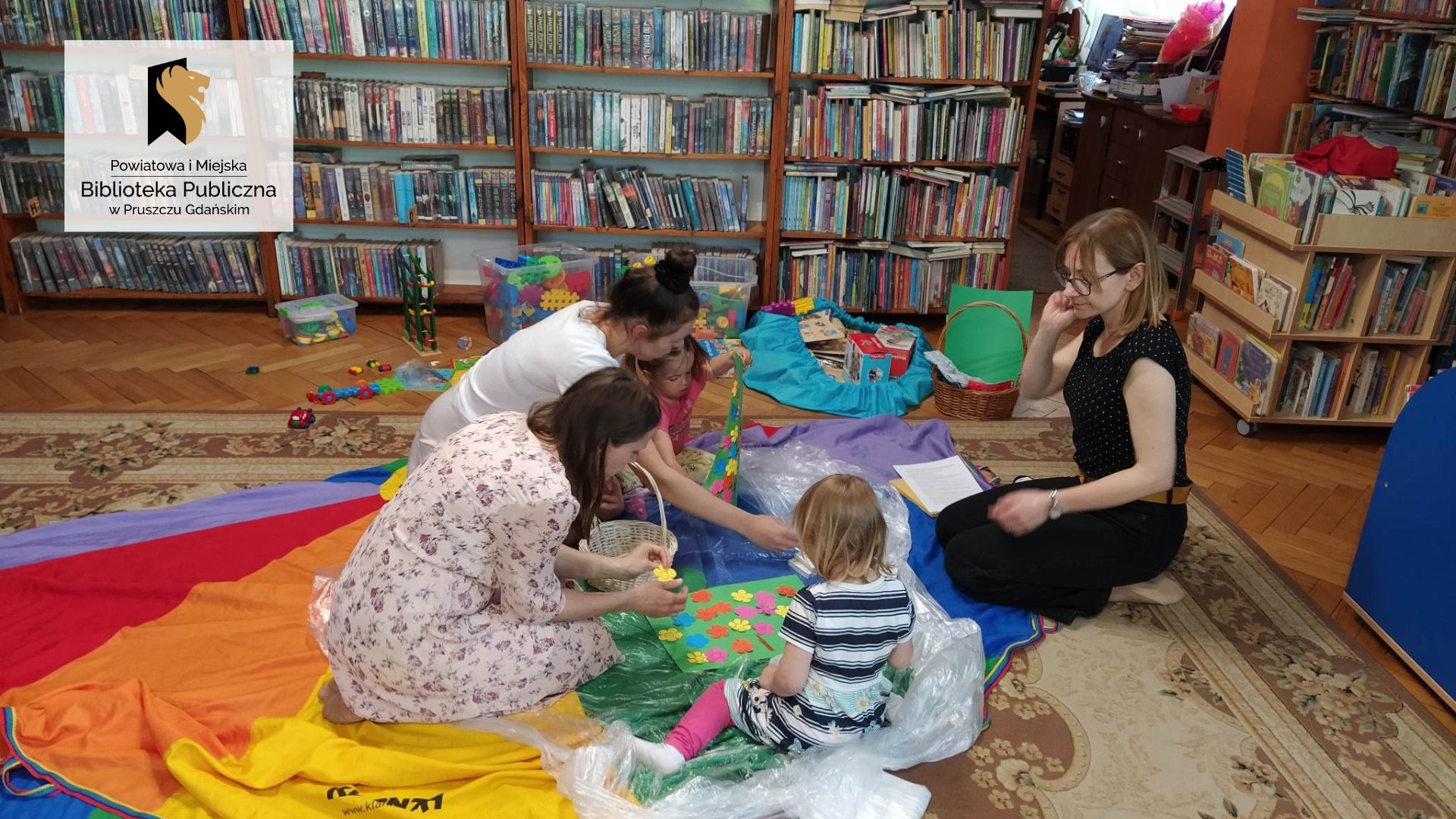 Dwie dziewczynki wraz z mamami i panią bibliotekarką siedzą na podłodze i przyklejają kolorowe kwiatki do kartki. Za nimi zabawki i regały z książkami.