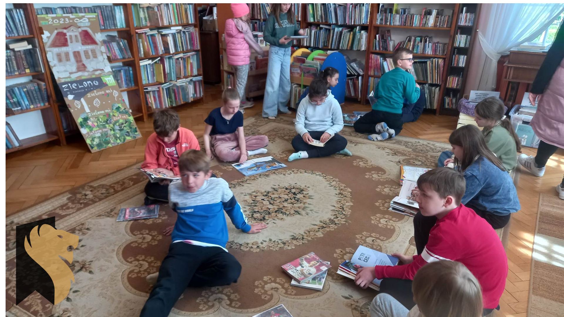 Dzieci siedzą na dywanie i oglądają książki. W tle regały z książkami.