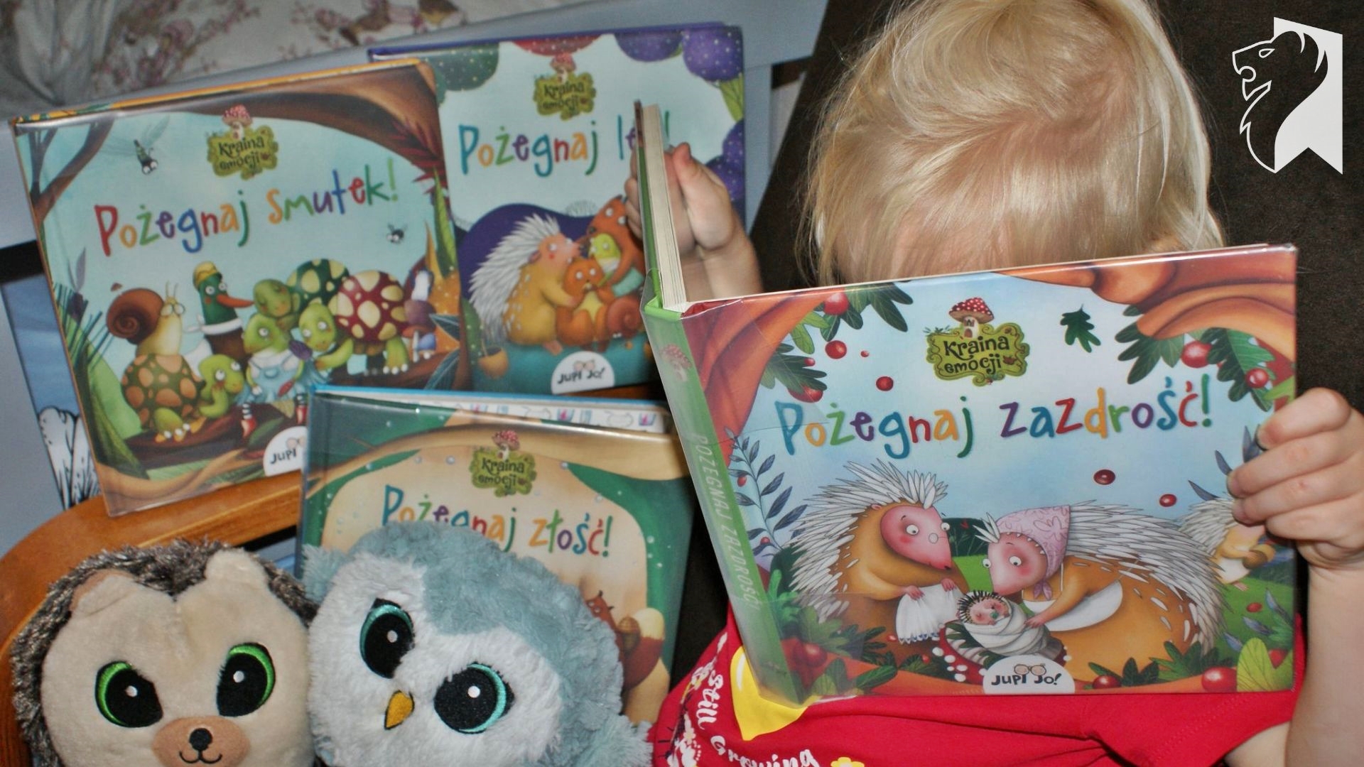 Dziecko trzyma w rękach książkę pt.: Pożegnaj zazdrość. Z lewej strony leżą jeszcze 3 książki tej serii: Pożegnaj smutek, Pożegnaj zazdrość, Pożegnaj lęk. W lewym, dolnym rogu, 2 pluszaki.