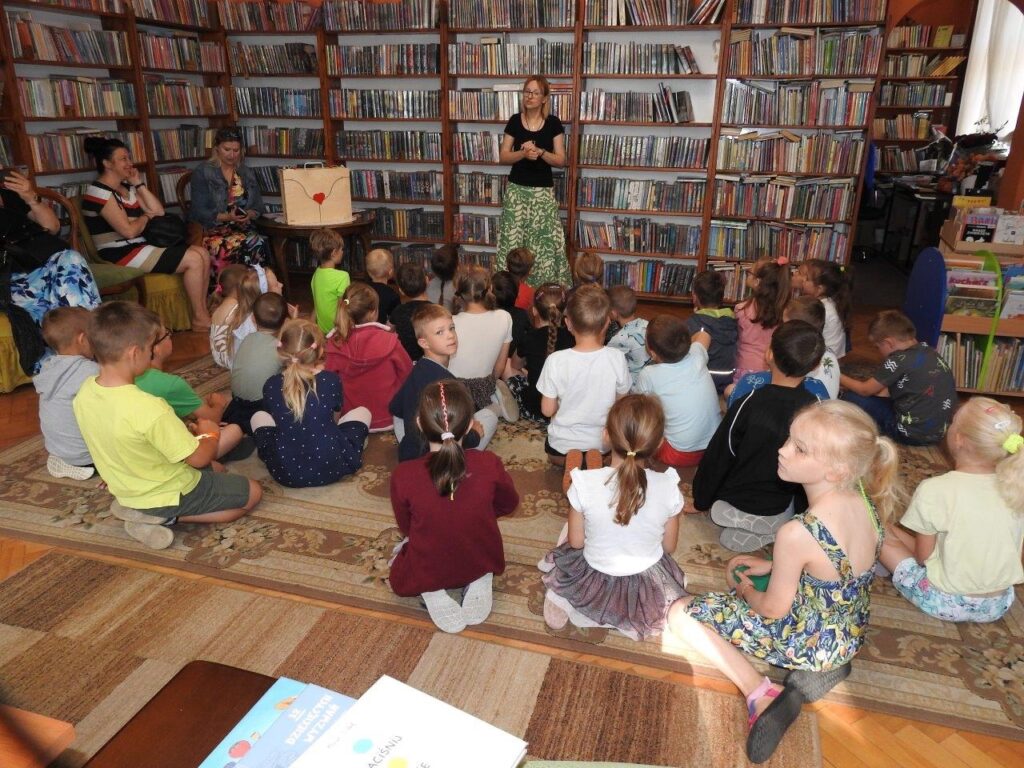 Dzieci siedzą na podłodze. Przed nimi stoi pani bibliotekarka i mówi. Po lewej siedzą nauczycielki. W tle regały z książkami.