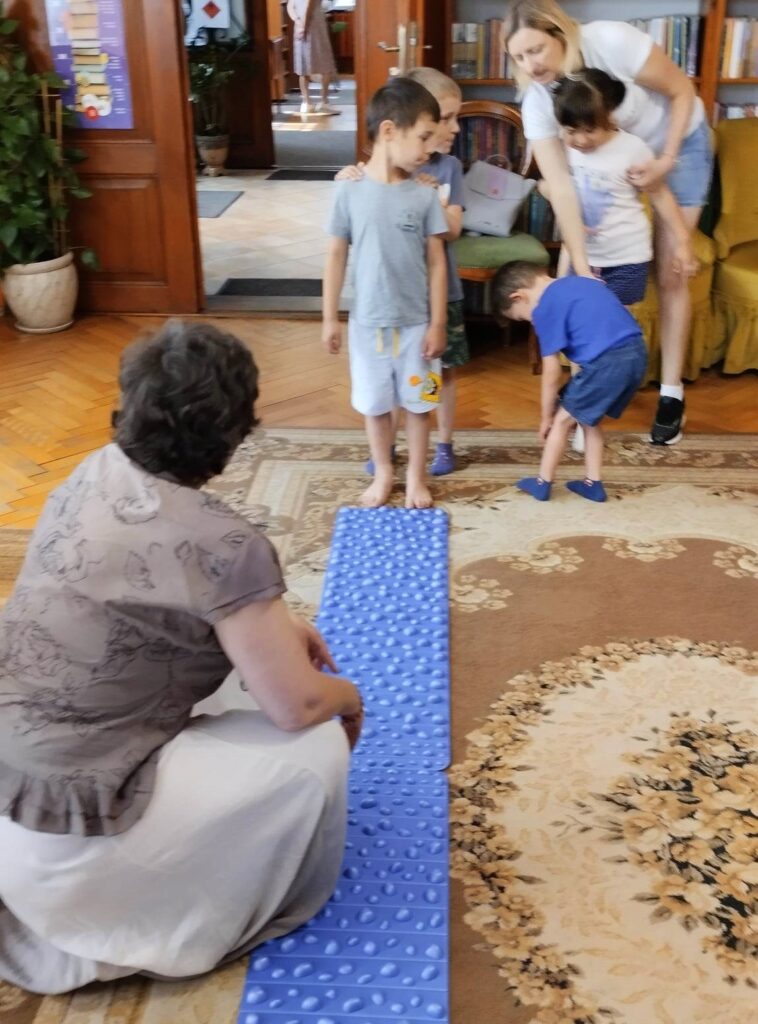 Na dywanie lezy niebieska mata sensoryczna. Przy niej kuca Pani Ania. Z przodu maty stoją dzieci i pani.