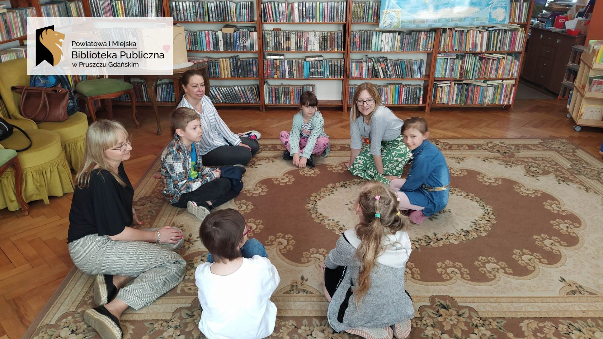 1. Dzieci wraz z paniami siedzą w okręgu na podłodze. W tle regały z książkami.